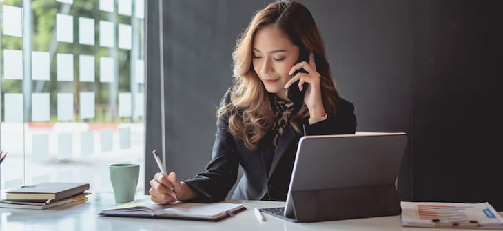 jeune femme au téléphone qui écrit dans un carnet devant son ordinateur portable