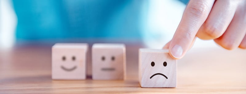 cube en bois avec un visage mécontent mi en avant par une main, en arrière plan 2 autres bloc en bois ont un visage souriant et neutre